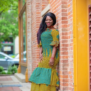 May include: A woman wearing a yellow and green patterned dress with a ruffled hem. She is standing next to a brick wall with a yellow door.