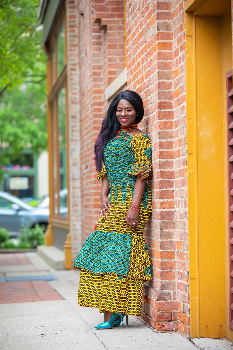 May include: A woman wearing a yellow and green patterned dress with a ruffled hem. She is standing next to a brick wall with a yellow door.