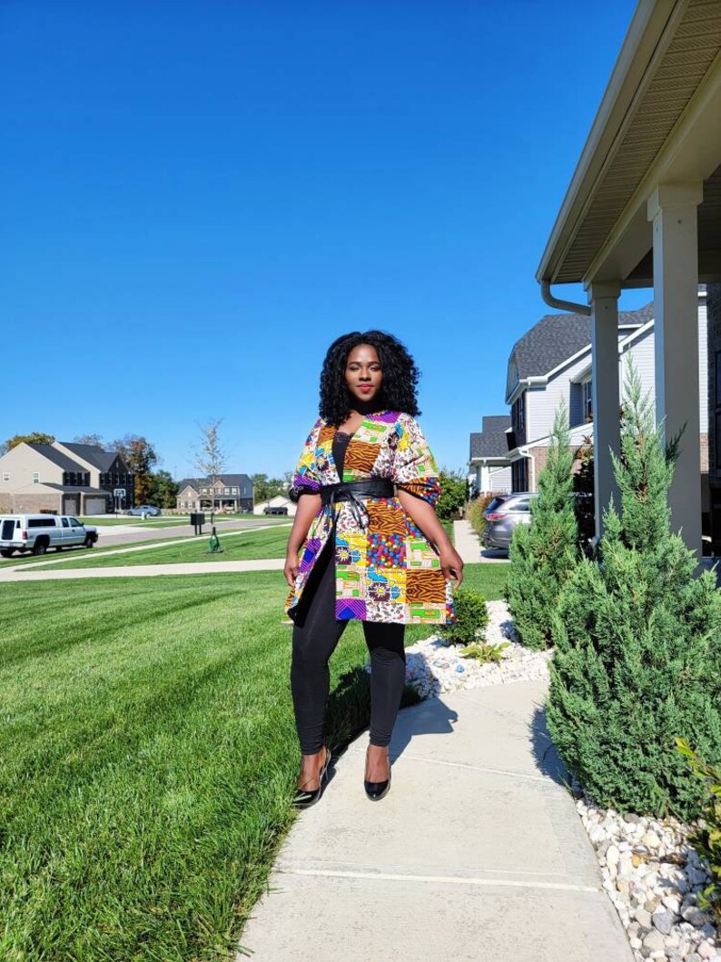 May include: A vibrant patchwork jacket, featuring a mix of patterns and colours, worn with a black belt and leggings. The jacket includes orange, yellow, blue, and purple. The outfit is completed with black heels.