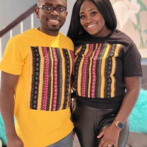 May include: A couple wearing matching black and yellow t-shirts with a colorful patterned design. The shirts have a black background with yellow and red stripes and a variety of geometric patterns.