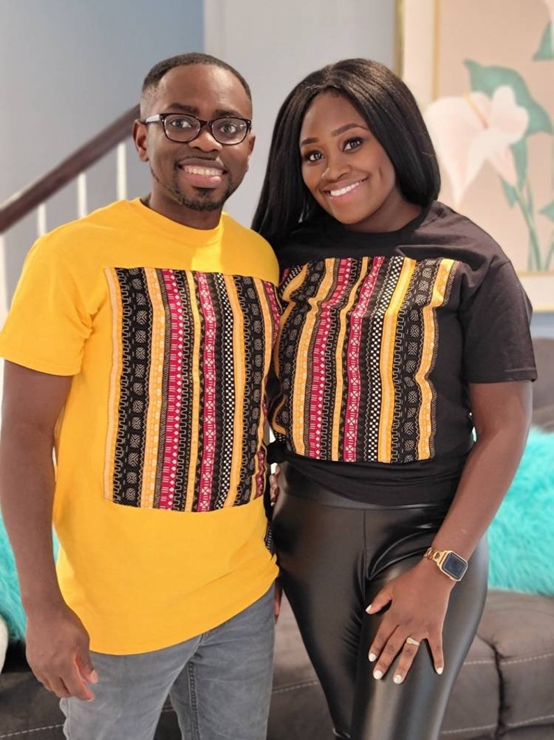 May include: A couple wearing matching black and yellow t-shirts with a colorful patterned design. The shirts have a black background with yellow and red stripes and a variety of geometric patterns.