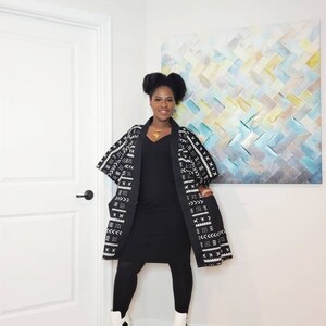 May include: A woman wearing a black dress and black leggings with a long black and white patterned cardigan. She is wearing white ankle boots and a gold necklace.