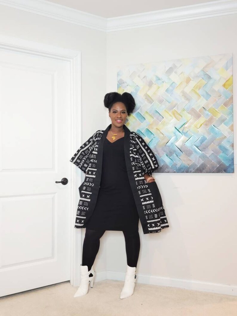 May include: A woman wearing a black dress and black leggings with a long black and white patterned cardigan. She is wearing white ankle boots and a gold necklace.