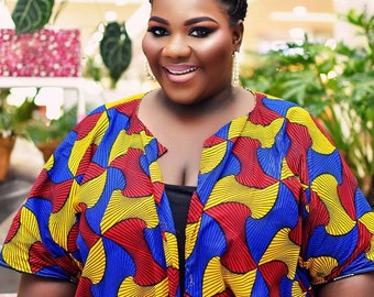 May include: A woman with a bright smile is wearing a blue, red, and yellow patterned shirt with a black undershirt. She has her hair in a braided bun and is wearing blue sunglasses.