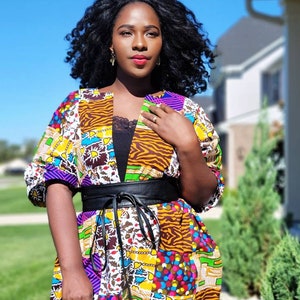 May include: A vibrant patchwork kimono jacket with a black belt. The jacket showcases diverse patterns, including animal prints and floral motifs, in yellow, purple, and brown hues. The person is wearing a black top underneath.