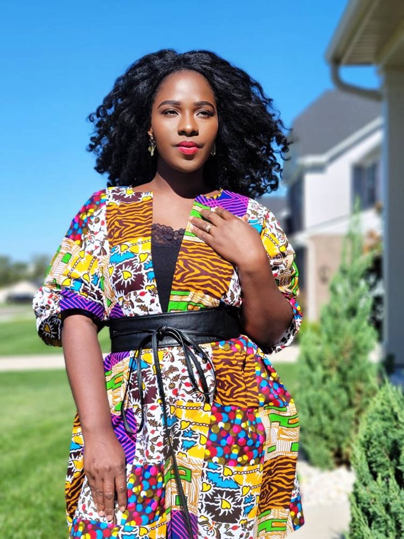 May include: A vibrant patchwork kimono jacket with a black belt. The jacket showcases diverse patterns, including animal prints and floral motifs, in yellow, purple, and brown hues. The person is wearing a black top underneath.