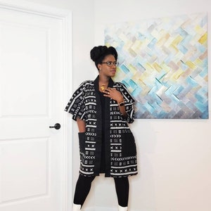 May include: A woman wearing a black and white patterned kimono with a black long-sleeved shirt and black leggings. She is wearing white ankle boots and has her hair in a bun. She is standing in front of a white wall with a painting behind her.