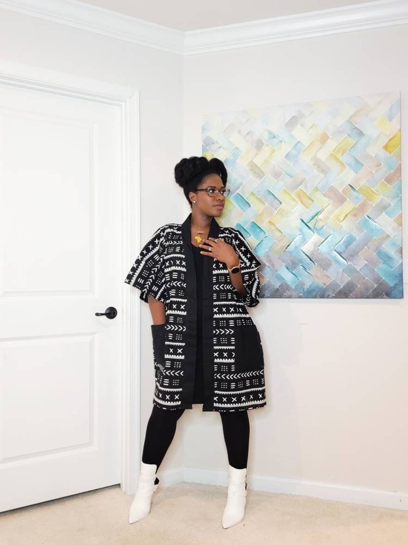 May include: A woman wearing a black and white patterned kimono with a black long-sleeved shirt and black leggings. She is wearing white ankle boots and has her hair in a bun. She is standing in front of a white wall with a painting behind her.