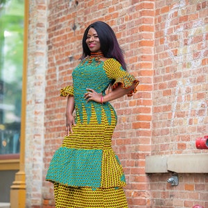 May include: A woman wearing a long, yellow and blue patterned dress with a ruffled hem. She is standing in front of a brick wall and has her hand on her hip.
