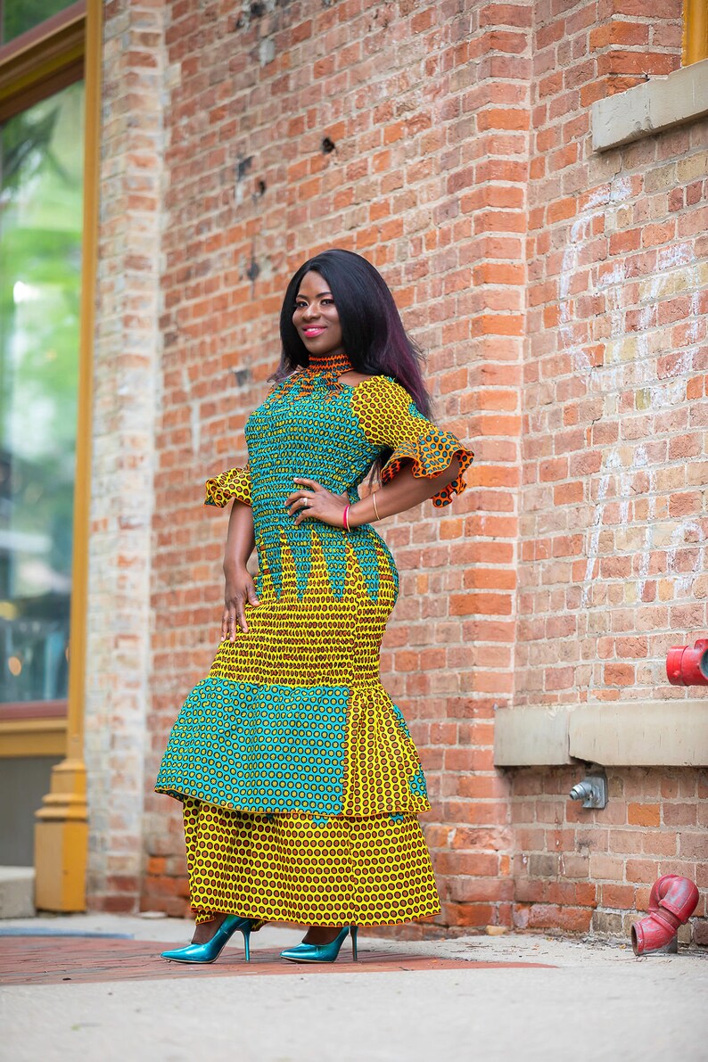 May include: A woman wearing a long, yellow and blue patterned dress with a ruffled hem. She is standing in front of a brick wall and has her hand on her hip.