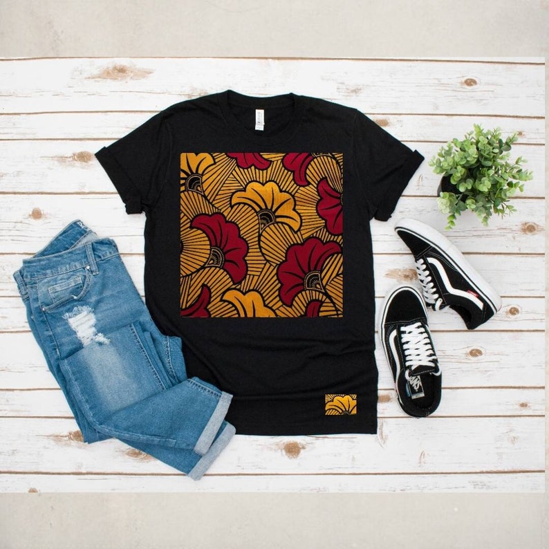 May include: Black t-shirt with a square print of a yellow and red floral pattern. The shirt is on a white wooden surface with a pair of blue jeans and black sneakers.