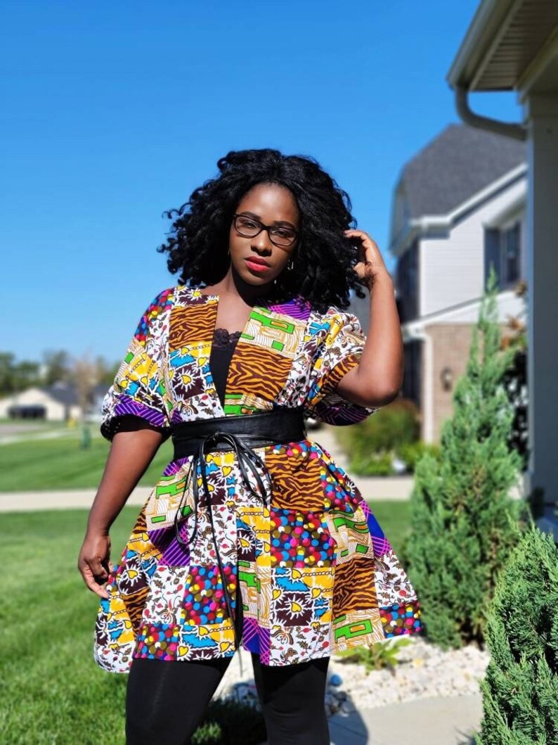 May include: A vibrant patchwork kimono-style top with a black leather belt. The top showcases diverse patterns, including animal print, geometric shapes, and floral designs. It has short sleeves and is worn over a black top and leggings.