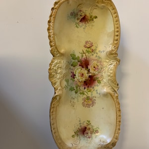 May include: A vintage, cream-colored serving dish with gold accents and floral designs. The dish has two handles and is perfect for serving snacks or appetizers.