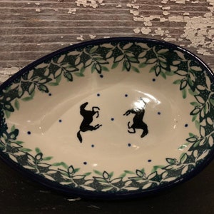 May include: White ceramic spoon rest with a dark blue rim and a decorative leaf pattern in green. Two black horse silhouettes are featured in the center. The spoon rest has a unique teardrop shape.