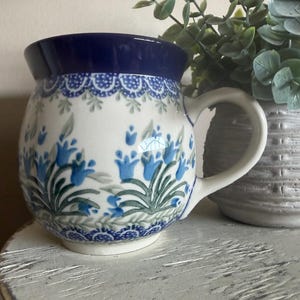 May include: A round ceramic mug with a dark blue rim and a white base featuring a hand-painted pattern of blue bell-shaped flowers with green leaves. The mug has a curved handle and a wide, bulbous body, showcasing a decorative scalloped border.