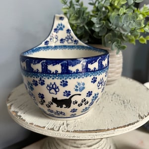 May include: A blue and white ceramic soup bowl with a paw print design and a black cat silhouette. The bowl has a handle and is sitting on a white pedestal.