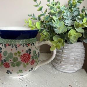 May include: A white ceramic mug with a blue rim and a floral pattern featuring green leaves and red clover flowers. The mug has a rounded body and a handle. It is sitting on a white surface next to a potted plant.