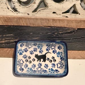 May include: A rectangular ceramic trinket dish with a cream-colored base, featuring a dark blue border and a repeating pattern of blue paw prints. A solid black silhouette of a cat is positioned in the center, surrounded by smaller black paw prints.