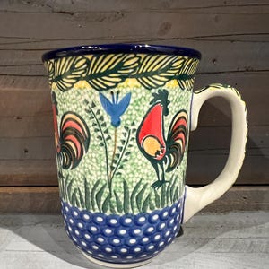 May include: A ceramic mug with a blue, green, and yellow rooster design. The mug has a white handle and a polka dot pattern around the bottom.