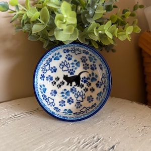 May include: A small, round ceramic bowl with a blue rim and a white interior. The bowl is decorated with blue paw prints and a black cat silhouette. The bowl is likely for a pet.