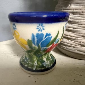 May include: A ceramic egg cup featuring a cream-colored base with a hand-painted floral pattern in shades of blue, yellow, and red. The cup has a thick, dark blue rim and a flared pedestal base, decorated with green leaves and small blue flowers.