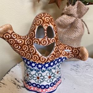 May include: A ceramic ghost-shaped candle holder with a smiling face and outstretched arms. The top portion features a brown swirl pattern, while the base has a floral design with blue, white, and coral accents. A unique Halloween decoration.