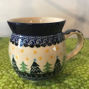 May include: A ceramic mug with a rounded shape and a dark blue rim. The mug features a festive design with green Christmas trees, yellow stars, and blue accents. The handle is also decorated with blue patterns.