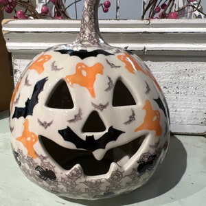 May include: A ceramic pumpkin figurine featuring a cream-colored base decorated with orange ghost silhouettes, black bats, and grey spider web patterns. The pumpkin has cut-out eyes, a nose, and a mouth, with a textured stem at the top.