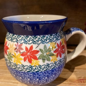 May include: A ceramic mug with a round body and a white handle. The mug features a blue rim and a pattern of colorful leaves in red, yellow, and green. The base of the mug has a blue floral design.