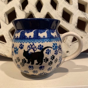 May include: A ceramic mug with a navy blue rim and handle. The mug features a white and blue cat paw print design, with a black cat silhouette. The mug has a rounded shape and a white interior.