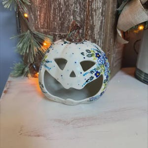 May include: A white ceramic pumpkin with a carved face and floral designs in blue and green. The pumpkin has a brown stem and is likely used as a Halloween decoration or candle holder. The pumpkin is sitting on a white surface.