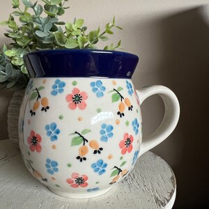 May include: A white ceramic mug with a blue rim and a floral pattern featuring pink, blue, and orange flowers. The mug has a rounded body and a handle.