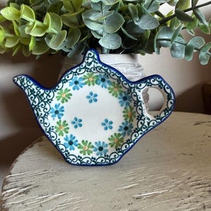 May include: A ceramic teapot-shaped tea bag holder with a white base and a blue rim. The holder is decorated with blue and green floral patterns. The handle is a small, open circle. The background includes a vase with green foliage.