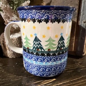 May include: A blue and white ceramic mug with a winter scene design. The mug features a repeating pattern of stylized pine trees, stars, and swirls. The mug has a handle and a wide rim.