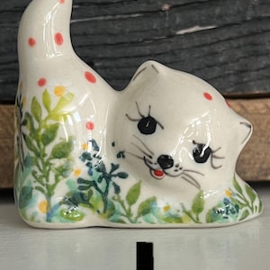 May include: A ceramic cat figurine in a reclining pose, painted with a cream base and adorned with red polka dots. The cat is decorated with green leaves, blue and yellow flowers, and black eyes. The figurine has a raised tail.