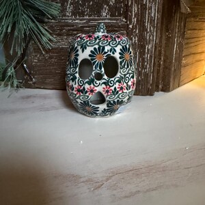 May include: A ceramic pumpkin-shaped candle holder with a floral pattern in green, orange, and red. The pumpkin has three eye-shaped cutouts and a small stem. The background includes a wooden door and a sprig of greenery.