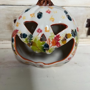 May include: White ceramic pumpkin with a carved jack-o'-lantern face. The pumpkin is decorated with a fall leaf and acorn pattern in shades of orange, red, green, and brown.