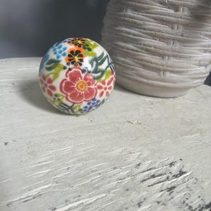 May include: A spherical ceramic knob featuring a hand-painted floral pattern with red, orange, blue, and green blossoms and leaves on a white background. The surface is glossy and smooth, showcasing a detailed botanical design across the entire round shape.