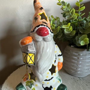 May include: A ceramic Halloween gnome figurine. The gnome wears an orange hat with black spiders and holds a lantern. The gnome's body is white with cut-out stars and is decorated with bats, pumpkins, and a black cat.
