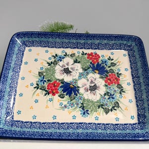 May include: Rectangular ceramic serving platter with a floral design. The center features a bouquet of white, red, and blue flowers on a cream background. The border is a dark blue with a decorative pattern. The platter is approximately 12 inches long and 8 inches wide.