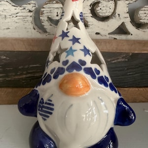 May include: A ceramic gnome figurine featuring a tall, pointed hat decorated with blue and red stars, hearts, and a flag-patterned heart. The gnome has a smooth orange nose, a white beard, and dark blue boots and mittens, with star-shaped cutouts.