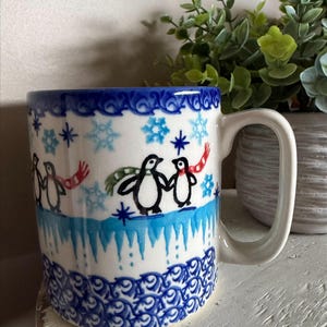 May include: White ceramic mug with a blue rim and handle, featuring a winter scene with penguins wearing scarves and snowflakes. The mug has a blue and white patterned design at the base and rim.