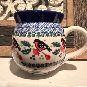 May include: A ceramic mug with a rounded shape, featuring a dark blue rim and a white body decorated with a pattern of red birds, green leaves, and red berries. The handle is white, and the mug has a capacity of approximately 12 ounces.