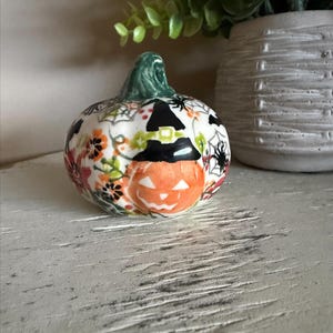 May include: A ceramic pumpkin figurine with a green stem, featuring a jack-o'-lantern face and a witch's hat. The pumpkin is decorated with floral and spiderweb patterns in orange, red, and black, perfect for Halloween decor.