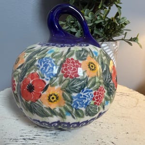 May include: A ceramic pumpkin-shaped decorative piece featuring a glossy dark blue stem and a cream-colored body adorned with a vibrant floral pattern of red, orange, and blue flowers surrounded by green leaves and a dark blue scalloped border.