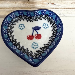 May include: Heart-shaped ceramic dish with a blue rim and a white interior. The interior features a cherry design with red cherries and a blue stem, surrounded by blue floral and leaf patterns. The dish is approximately 4 inches wide.