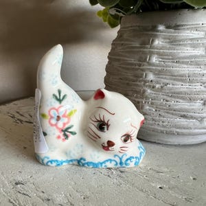May include: A white ceramic cat figurine with blue and pink floral details. The cat is curled up with its tail raised and has a painted face with black eyes and a pink nose.