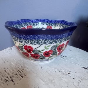 May include: A ceramic bowl with a scalloped rim, featuring a hand-painted pattern of red pansies with green leaves on a white background. The upper border is decorated with a dark blue band containing a repeating white and green floral motif.