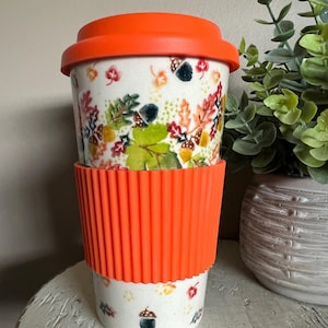May include: A white travel mug with an orange lid and a matching orange silicone sleeve. The mug is decorated with colorful autumn leaves and acorns. The mug is designed for hot or cold beverages.