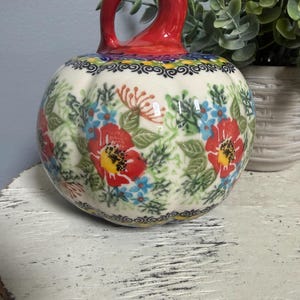 May include: A ceramic pumpkin-shaped decorative piece with a glossy red stem. The body is cream-colored and features a hand-painted floral pattern with red poppies, small blue flowers, green leaves, and a black decorative border around the top.